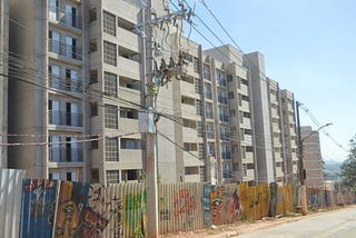 Prédio residencial de vários andares com fachada cinza claro, localizado atrás de uma cerca de metal colorida com grafites. Em primeiro plano, há um poste de eletricidade com fios e transformadores suspensos que cruzam a imagem. À direita, uma rua asfaltada acompanha a lateral do condomínio, que se estende ao fundo sob um céu azul e limpo.