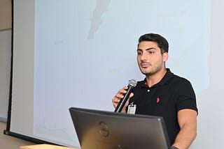 Close-up of Nazih with a microphone in front of a presentation screen.