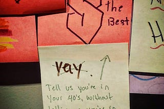 Pink sticky note which has a 90’s “S” written on it with words next to it stating “I’m the best”. Yellow sticky note under this has “yay” crossed out and an arrow pointing to sticky note above. Words under arrow state “Tell us you’re in your 40’s, without telling us you’re in your 40s.”