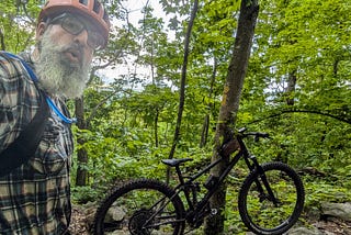 The author, a bearded man wearing a red helmet, glasses, a plaid shirt, and a hydration pack, takes a selfie in a dense, green forest. He appears slightly breathless or surprised. Beside him is a black mountain bike resting against a tree. The trail is rocky and surrounded by lush foliage, indicating a rugged and remote woodland setting.
