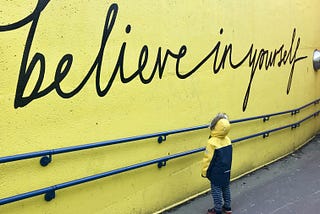 Child looks at a yellow wall with the words believe in yourself