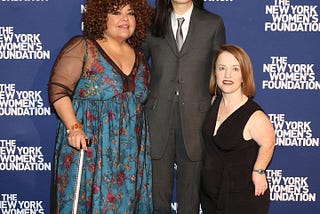 Three women standing against a blue and white backdrop reading “NY Women’s Foundation. One is an African and Indigenous ancestry with curly shoulder length hair and a dress that has sheer black sleeves and a blue flowered pattern. Next to her is an Korean American woman in a grey suit with a grey tie and a white shirt. Next to her is a white, achondroplastic dwarf woman with red hair and freckles wearing a Black sleeveless jumpsuit.