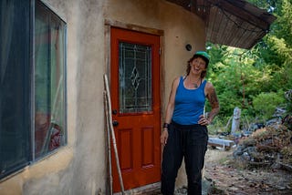 A smiling woman stands outside a house that has rough natural plaster walls and a red door.