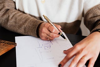 A person sketching something on pen and paper, while a laptop is right next to them