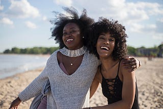Two beautiful black women embrace each other while smiling on the beach. Their afros blow in the wind as the sun kissed their skin.