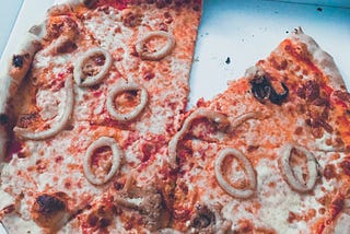 A pizza missing a slice in a box. The pizza is topped with seafood including calamari rings and tiny octopus.