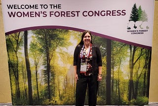 Candra Burns wearing business casual clothing in front of a sign saying Welcome to The Women’s Forest Congress with a forest background.