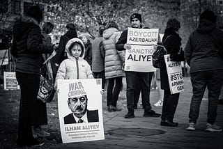 A child holding a sign with “War Criminal: Ilham Aliyev” and a man holding a sign with “Boycott Azerbaijan / Sanction Aliyev”
