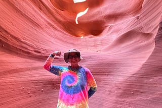 CC appears in a rainbow tie dyed long sleeve hoodie and a tan hiking hat standing in front of one of the orange/red canyon walls of lower antelope canyon and doing the classic colorectal cancer strong arm selfie.