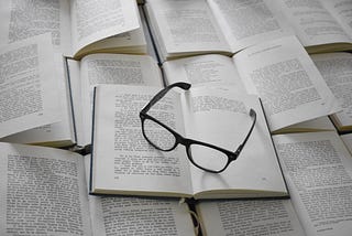 Many books, one of top of each other with glasses on top.