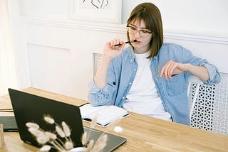 A woman writing at her desk, looking pensively at the laptop with a pen pressed at her lips and a notebook at her side.