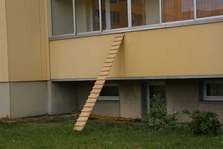 A cat ladder leans against a balcony wall of a Soviet Era style apartment building in Lithuania. The ladder is a board about 4 meters in length with about 15 small slats attached horizontally to act as stairs or steps on the ladder.