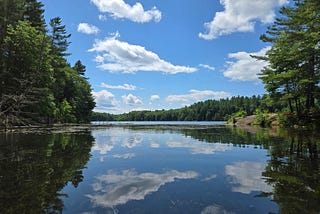 A gorgeous day and the blue sky, filled with puffy clouds, is reflected by the smooth surface of a lake. Conifer trees rise on the banks and on the distant hills. It is peaceful.