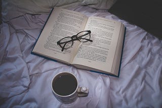 A book set on top a bed with a pair of folded reading glasses on the book. Nearby is a laptop and a cup of coffee.