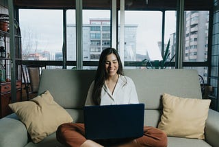 Woman sits on couch while looking at her laptop and smiling. There is a large window behind her with two apartment blocks in the background.