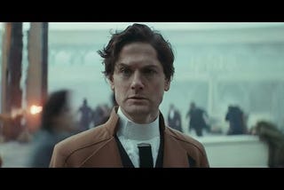 image of the character Syril Karn from the TV show Andor. He is standing in a plaza, looking bewildered, as a fight between fascists and rebels erupts all around him