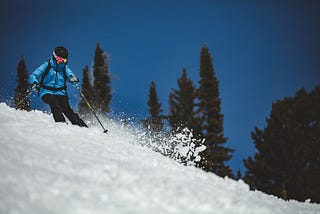 Skier in blue ski jacket, black helmet and ski pants, and multicolored ski goggles skiing down a slope with fresh snow against green evergreens and a dark blue sky.