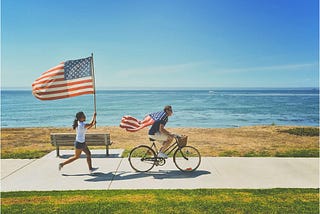 Woman holding American flag high while chasing bike ridden by a man wearing the American flag over his shoulders.