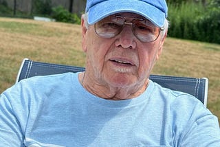 This photo shows an older man dressed in a blue T-shirt and cap sitting in a lawn chair. The background is a yard, and it is summertime.