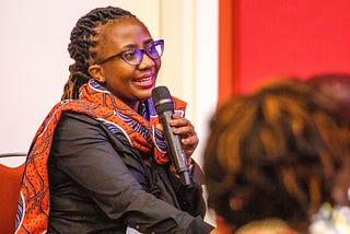 A Kenyan woman with braids and glasses speaks into a microphone