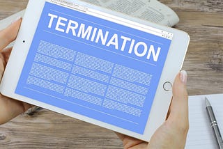 Hands of a person holding a white iPad with a blue screen with white font that reads “TERMINATION” with three columns of illegible text below.