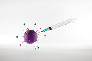A hypodermic needle injecting a purple ball with colored pins representing a virus.