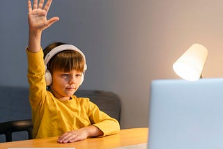 Young boy in a yellow sweater wearing headphones sitting in front of a laptop at a desk raises his hand during his homeschooling lesson