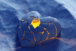 A stone heart, cracked with light showing through rests in silver sand.