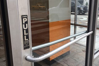 A glass door with bars on the front and back, seen from a viewpoint outside a building. A vertical sign reading “PULL” is installed over the pull bar. Someone exiting the building would encounter the pull bar, but would have to push.