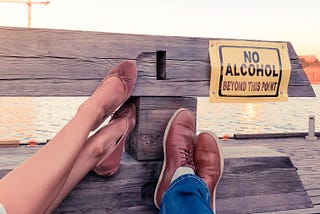 Two people rest their feet against a wooden fence displaying a yellow “No Alcohol Beyond This Point” sign. The sun sets over water in the background.