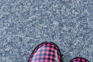 The toes of two buffalo plaid rainboots peak into a frame of distressed grey concrete.