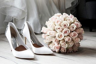 A pair of white wedding shoes on a hardwood floor next to a light pink rose bouquet.