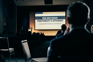 View from inside a conference audience. An out-of-focus attendee sits in the foreground, watching a distant leader on stage present a slide reading “OUR NEW FOCUS: PROFITABILITY & EFFICIENCY.” The auditorium is dim; the screen glows against silhouetted heads.
