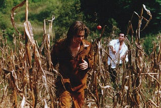 A man dressed like Tom Sawyer runs through a field laughing, as a tomato whizzes through the air, hurled by another man, seen in the distance, wearing a white button up shirt.