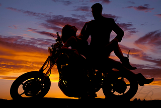 Silhouette of a woman lying back on a motorcycle as a man prepares to straddle her.