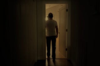 The silhouette of a man entering a dimly lit room from a darkened hallway.