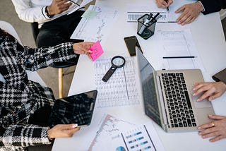 People working at a desk, with papers, laptops, and tablets all over. People are writing thigns down, looking at computers, and more