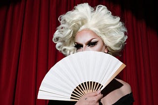 A drag queen holding a fan in front of her face.