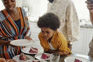 young brown skin boy in center of photo with mustard colored shirt starring at a plate with strawberry cake. a black lady on his right with a blue and orange dress on holding a plate. a man with a white shirt is behind boy, face out of frame