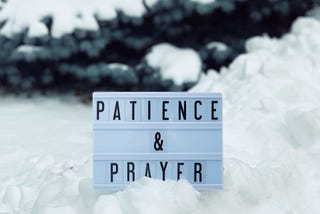 A board with text patience and prayer on the snow with a tree in the background