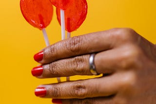 Woman’s hand holding 3 red heart-shaped lollipops like a bouquet, against an orangey-yellow background.