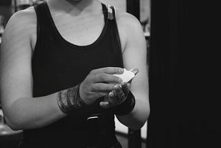 a black and white close up photo of a weightlifter in a singlet chalking their hands. the photo crops the athlete from neck to waist and focuses on their hand. the right side of the photo is plain black, being the curtain between the back warm up area and the audience.