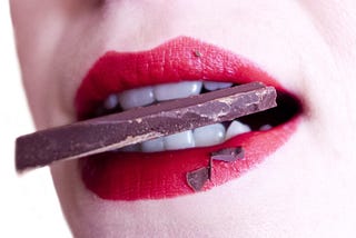 Closeup of a pale-skinned woman with red-lipsticked lips holding a flat piece of chocolate between her very white teeth. Only her lower face is shown.