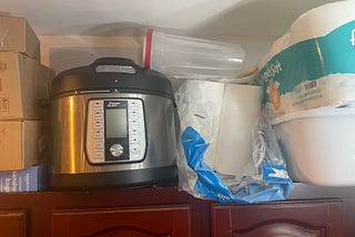 rice cooker, paper products and a plastic container all stored on top of a cabinet