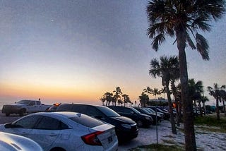 author’s white honda civic backed into a parking spot in the sand in a parking lot on Florida’s Gulf Coast, with palm trees and the sunset in the background