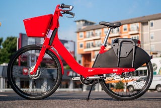 A bright orange-red E-bike on a plaza with a multi-story building and a blue sky in the background