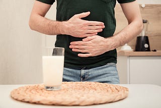 A lactose-intolerant man in a black t-shirt rubs his stomach in front of a glass of milk.