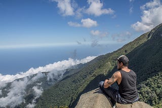 Photo of a man perched on a mountain looking out over an ocean.