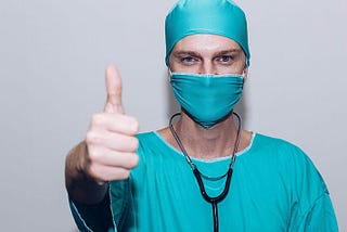 A man wearing blue scrubs, a mask and head covering, with a stethoscope,  is doing a thumbs up.
