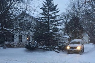 Victorian-style house photographed after a day of snow and cold. Light in the window, and a snow-covered landscape, and bare trees.